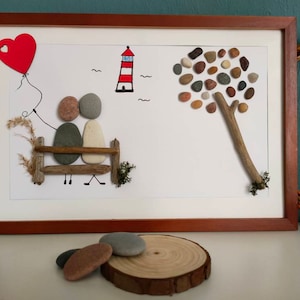 Könnte beinhalten: Ein gerahmtes Kunstwerk mit einem Paar aus bemalten Steinen, das auf einer Treibholzbank sitzt und einen roten Herzluftballon hält. Das Kunstwerk enthält auch einen bemalten Leuchtturm, einen Baum aus Steinen und ein kleines Herz.