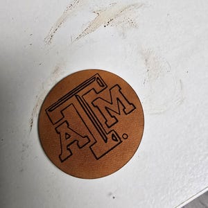 May include: A brown leather coaster with the letters "A&M" in black outlined lettering.