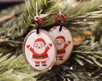Puede incluir: Un par de pendientes ovalados con fondo transparente y una imagen de dibujos animados de Papá Noel con un traje rojo y barba blanca. Los pendientes cuelgan de ganchos plateados con cuentas rojas y negras.