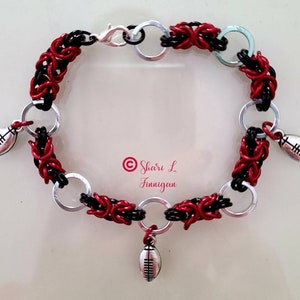 Könnte beinhalten: Ein rot-schwarz-silbernes Kettenarmband mit drei silbernen Fussballanhängern. Das Armband ist aus geflochtenem Draht gefertigt und hat einen Karabinerverschluss.