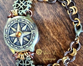 Może przedstawiać: Bransoletka w stylu steampunk z zawieszką w kształcie kompasu, złotymi i srebrnymi ogniwami łańcucha oraz dwoma zawieszkami w kształcie zębatki. Tarcza kompasu ma złotą gwiazdę z żółtym kamieniem w środku.