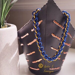 Peut inclure: Un bracelet en maille de chaîne bleu et or. Le bracelet est composé de petits anneaux métalliques qui s'emboîtent pour créer un motif de maille de chaîne. Le bracelet est sur un fond noir.