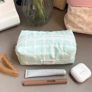 May include: A rectangular quilted pouch featuring a light mint green and white plaid pattern with a white zip and a small fabric label reading by annabel. The pouch is accompanied by a light blue tube, a brown pen, a white earbud case, and a hair clip.