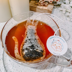 May include: A clear glass teacup with a cat-shaped tea bag and a festive tag that reads "Meowry Christmas!"  The tea bag is partially submerged in the amber-colored tea. The teacup is on a white countertop with a gold decorative tree and candles in the background.