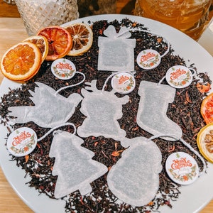 May include: A white plate filled with loose leaf tea and Christmas-themed tea bags. The tea bags are shaped like a Christmas tree, a stocking, a reindeer, a snowman, a gift box, and a bird. There are also dried orange slices and round tags that say "Merry Christmas".