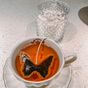 May include: A white teacup and saucer with a gold trim, filled with tea and a butterfly-shaped tea bag. A clear glass sugar bowl with a lid sits in the background.