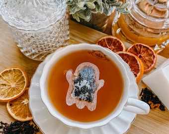 Puede incluir: Una taza de té blanca con una bolsita de té con forma de fantasma en una taza de té. La taza de té está sobre un platillo blanco con un borde dorado. Hay rodajas de naranja secas y té suelto sobre una superficie de madera.
