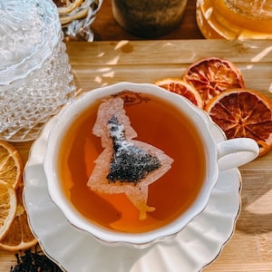 May include: A white teacup and saucer with a tea bag shaped like Alice in Wonderland. The tea bag is floating in the brown tea. The teacup and saucer are on a wooden surface with dried orange slices.