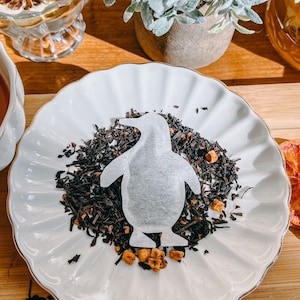 May include: A white tea bag shaped like a penguin on a white plate with loose leaf black tea and orange pieces.