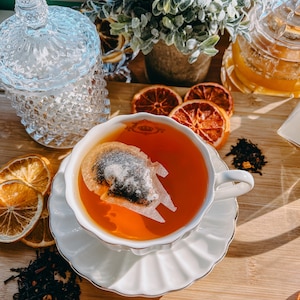 May include: A white teacup and saucer with a gold rim, filled with orange tea and a tea bag shaped like a spaceship. The teacup is on a wooden surface with dried orange slices and loose leaf tea.
