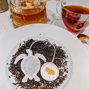 May include: A white tea bag shaped like a turtle with the text "I shell always love you" and a small turtle illustration on a white circle. The tea bag is surrounded by loose tea leaves on a white plate.