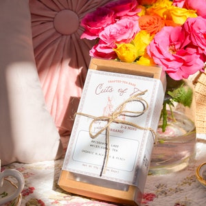 K&ouml;nnte beinhalten: Eine Schachtel Cuts of Joy Teebeutel mit einem braunen und wei&szlig;en Etikett. Das Etikett tr&auml;gt die Aufschrift "Crafted Tea Bags" und "Cuts of Joy". Die Schachtel ist mit Schnur gebunden und hat eine kleine Hasenillustration. Die Schachtel steht auf einer wei&szlig;en und rosa floral gemusterten Oberfl&auml;che.