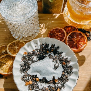May include: A white tea bag sits in a white ceramic bowl filled with loose leaf black tea. Dried orange slices are scattered around the bowl on a wooden surface.