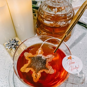 May include: A glass teacup with a red tea bag shaped like a snowflake. The teacup is on a white surface with a gold-colored saucer. The tea bag has the text "Happy Holidays!" on it.