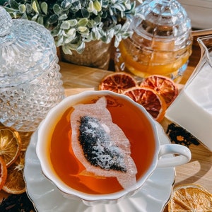 May include: A white teacup and saucer with a tea bag floating in a cup of amber tea. Dried orange slices and loose leaf tea are scattered on a wooden surface.