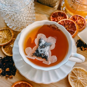 May include: A white teacup and saucer with a tea bag in the shape of a bear. The teacup is filled with brown tea. The saucer is decorated with a gold trim. The teacup and saucer are surrounded by dried orange slices and loose leaf tea.