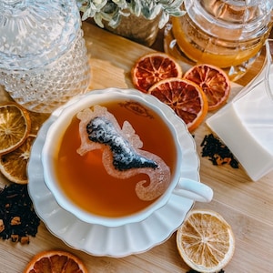 May include: A white teacup and saucer with a tea bag in the shape of a mermaid. The teacup is filled with amber-colored tea. Dried orange slices and loose leaf tea are scattered around the teacup on a wooden surface.