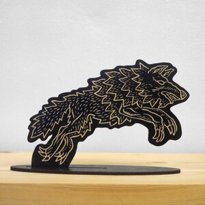 May include: A black wooden sculpture of a leaping wolf with detailed etched patterns. The wolf is mounted on a black base, creating a three-dimensional effect. The sculpture is displayed on a light-coloured wooden surface.