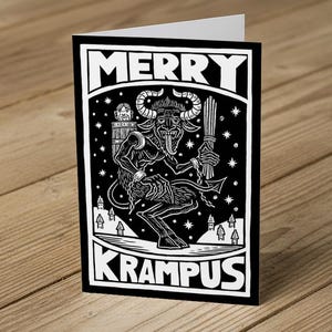 May include: A black and white greeting card with the text "MERRY KRAMPUS". The illustration depicts a horned demon holding a bundle of sticks, set against a backdrop of stars and small houses. The card is folded.
