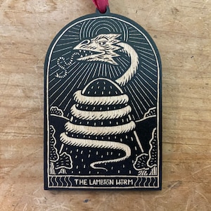 May include: A black wooden ornament with a white design of a dragon-like creature coiled around a hill. The text "THE LAMBTION WORM" is at the bottom of the ornament.