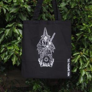 May include: Black tote bag featuring a white illustration of a witch stirring a cauldron. The witch is wearing a pointed hat and holding a wooden stick. The text "THE FOLKLORE BOX" is printed in white on the lower right corner of the bag.