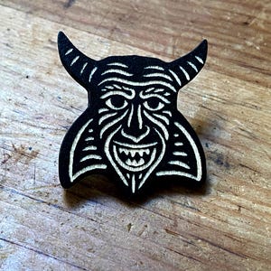 May include: A black wooden pin depicting a devil's face with horns, a wide grin, and detailed facial features. The design is carved with white accents, creating a contrast against the dark background. The pin is set against a wooden surface.