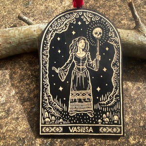 Puede incluir: Adorno de madera negro con una mujer sosteniendo un bastón con una calavera, rodeada por un diseño de bosque. La palabra "VASILISA" está en la parte inferior. El adorno tiene una cinta roja para colgar.