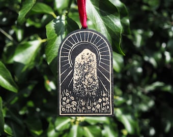 Puede incluir: Un adorno de madera negro con una imagen dorada de una piedra de pie, rodeada de hojas y un diseño de rayos de sol.
