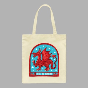 May include: A beige tote bag with a red dragon illustration inside a blue and white arched frame. The text "HERE BE DRAGONS" is printed below the frame.