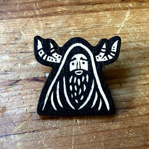 May include: A wooden pin featuring a black and white design of a bearded figure in a hooded cloak. The figure has a long beard and stylized wings or horns extending from the hood. The pin is set against a wooden surface.