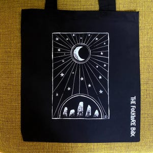 May include: Black tote bag with a white celestial design featuring a crescent moon, stars, and sunburst rays. The bottom of the design includes a curved line with several vertical shapes. The text "THE FOLKLORE BOX" is printed vertically on the right side.