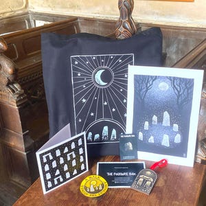 May include: A black tote bag with a white moon and stone circle print, a matching art print, a folded greeting card with a stone arch pattern, a circular yellow sticker reading A WALK AMONGST THE STONES, a small business card, and a wooden ornament.