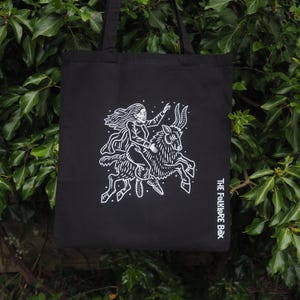 May include: Black tote bag featuring a white illustration of a person riding a horned animal, with flowing hair and outstretched arm. The text "THE FOLKLORE BOX" is printed in white on the lower right corner.