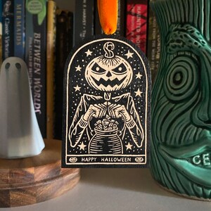 May include: A black wooden ornament with a white design of a pumpkin-headed figure holding a bag of candy. The ornament has a star-shaped background and the text "Happy Halloween".
