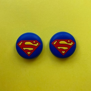 May include: Two blue round buttons, each featuring a red and yellow Superman logo.