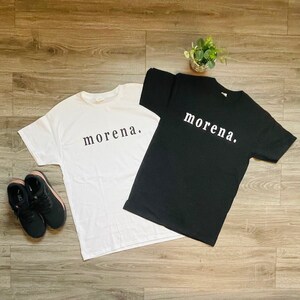 May include: Two t-shirts, one white and one black, each with the word "morena." printed in a simple, sans-serif font. The shirts are laid flat on a wooden surface, with a pair of black sneakers with rose gold accents and a small plant in the background.
