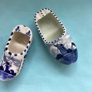 Könnte beinhalten: Zwei blaue und weiße Keramik-Aschenbecher in Form von holländischen Holzschuhen. Die Schuhe sind mit einer Windmühle und dem Wort "Amsterdam" verziert.