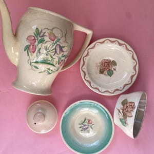 May include: A vintage cream-coloured tea set with floral patterns. The set includes a teapot, lid, two saucers, and a teacup. The teapot has a handle and spout. The saucers and teacup feature a rose design. The set is displayed on a pink surface.