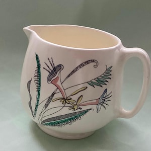 May include: Cream-coloured ceramic jug with a curved spout and handle. The jug is decorated with a floral design in shades of pink, green, and grey. The flowers have trumpet-like shapes and are surrounded by stylised leaves.