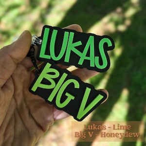 May include: A two-piece keychain set featuring black rectangular tags with raised lime green lettering. The top tag displays the name LUKAS, and the bottom tag displays the text BIG V. Both tags are connected by a black metal ring and clasp.