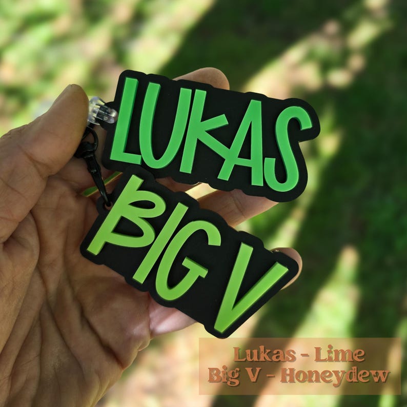 May include: A two-piece keychain set featuring black rectangular tags with raised lime green lettering. The top tag displays the name LUKAS, and the bottom tag displays the text BIG V. Both tags are connected by a black metal ring and clasp.
