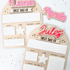May include: Wooden first day of school signs with the names "Lana", "Ayah", and "Jules" in pink. Each sign has space for date, age, things I love, and what I want to be. The signs are decorated with stars.