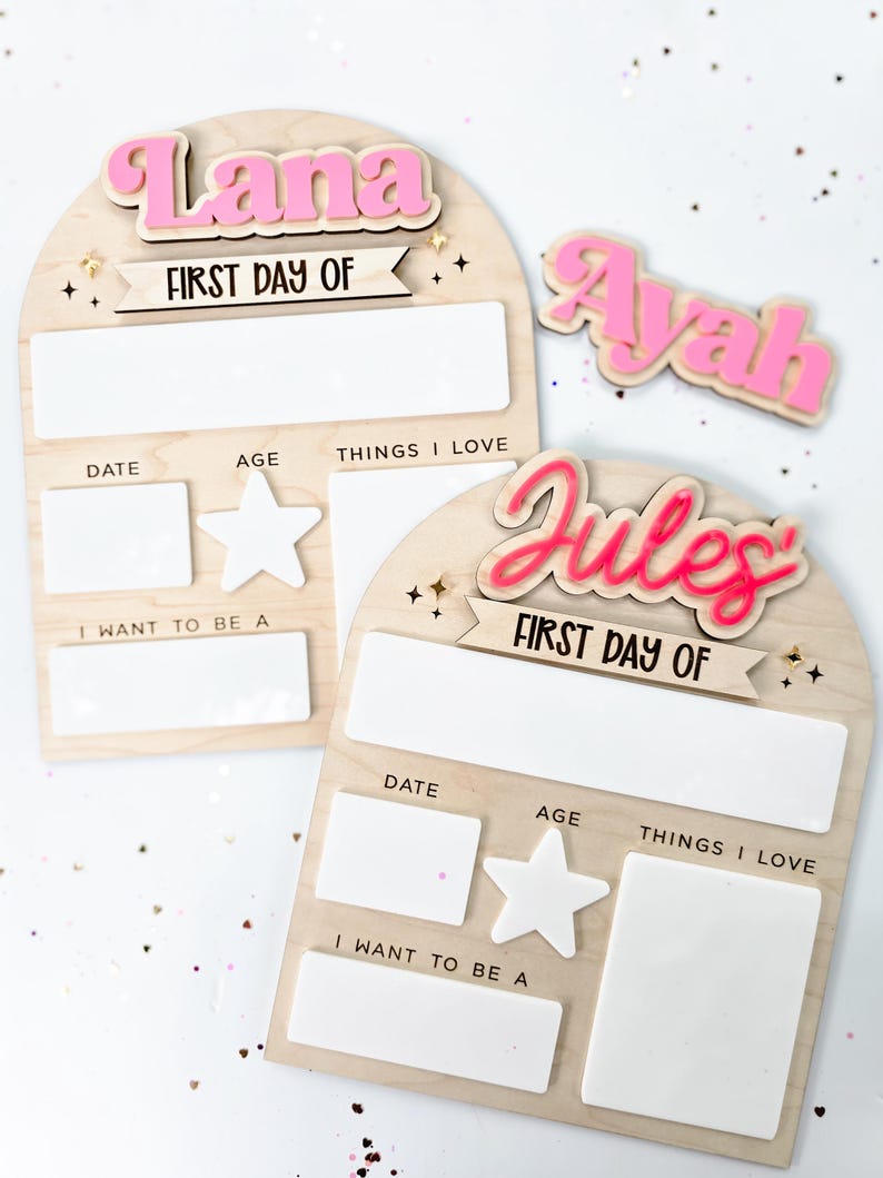 May include: Wooden first day of school signs with the names "Lana", "Ayah", and "Jules" in pink. Each sign has space for date, age, things I love, and what I want to be. The signs are decorated with stars.