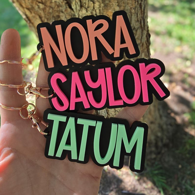 以下が含まれることがあります： ピーチ色の「NORA」、ホットピンク色の「SAYLOR」、ミントグリーン色の「TATUM」という名前が刻まれた、3つのカスタムキーホルダー。各名前は太いサンセリフ体で、黒い縁取りが施されており、ゴールドトーンの金属製ロブスタークラスプに取り付けられています。