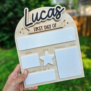 May include: Wooden sign with the name "Lucas" in black script, "FIRST DAY OF" on a banner, and blank spaces for details. Includes a star and starburst accents. A personalized back-to-school keepsake.