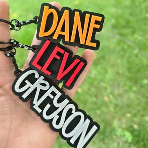 May include: Three rectangular keychains with black borders and black metal lobster clasps. The top keychain features the name DANE in orange, the middle keychain features the name LEVI in red, and the bottom keychain features the name GREYSON in white.