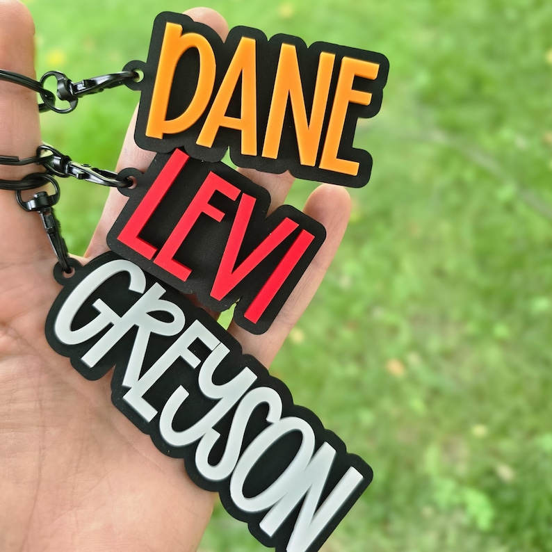 May include: Three rectangular keychains with black borders and black metal lobster clasps. The top keychain features the name DANE in orange, the middle keychain features the name LEVI in red, and the bottom keychain features the name GREYSON in white.