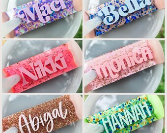 Puede incluir: Una colección de seis etiquetas rectangulares con diversos fondos brillantes y letras en relieve. Los nombres Maci, Bella, Nikki, Monica, Abigal y HANNAH se muestran en diferentes fuentes sobre superficies de resina con destellos.