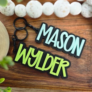 May include: Two custom keychains featuring black plastic bases with raised lettering. One keychain displays the name MASON in light blue, and the other displays the name WYLDER in lime green. Each keychain includes a black metal ring and clasp.