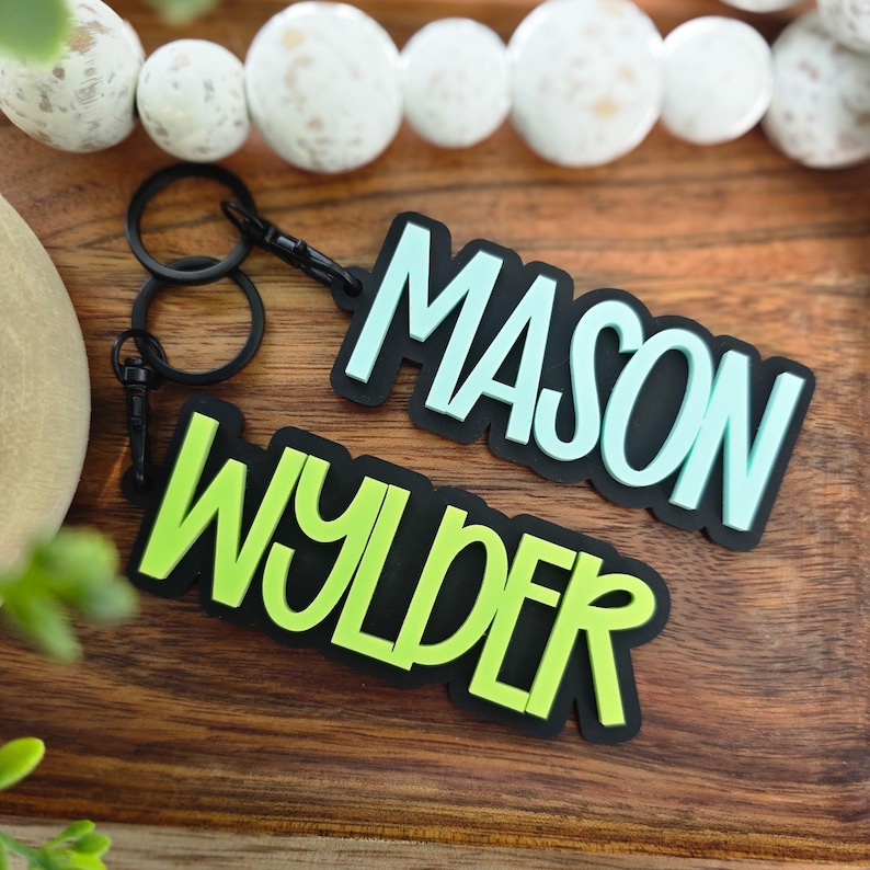 May include: Two custom keychains featuring black plastic bases with raised lettering. One keychain displays the name MASON in light blue, and the other displays the name WYLDER in lime green. Each keychain includes a black metal ring and clasp.